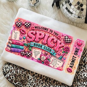 May include: White sweatshirt with a pink and black graphic that reads "WELCOME TO THE SPICY BOOK CLUB". The design includes hearts, cherries, roses, and a ticket that says "ADMIT ONE".