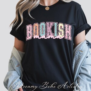 May include: Black t-shirt with the text "BOOKISH mama" in a colorful floral design. The word "BOOKISH" is in a large, blocky font, and "mama" is in a cursive font. The shirt is a casual style.