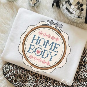 May include: A white sweatshirt with a cross-stitch design featuring the words "HOME BODY" in blue and a pink heart. The design is set within a brown embroidery hoop. The sweatshirt is folded over a leopard print fabric.