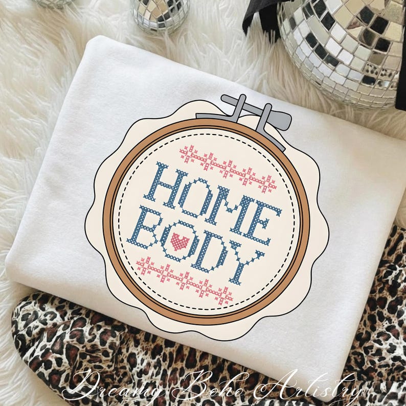 May include: A white sweatshirt with a cross-stitch design featuring the words "HOME BODY" in blue and a pink heart. The design is set within a brown embroidery hoop. The sweatshirt is folded over a leopard print fabric.