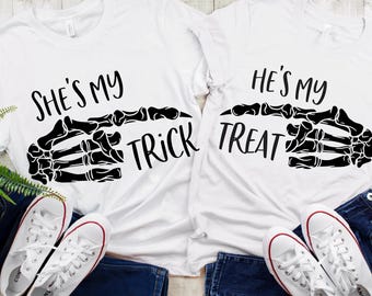 May include: Two white t-shirts with black skeleton hand graphics. One shirt says "SHE'S MY TRICK" and the other says "HE'S MY TREAT". White sneakers with red accents and blue jeans are visible.