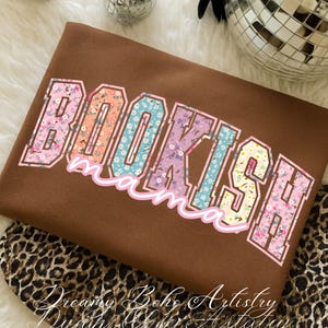 May include: A brown sweatshirt with the text "BOOKISH mama" in a floral design. The letters are outlined in white and filled with various floral patterns in pink, blue, orange, and purple. The sweatshirt is folded.