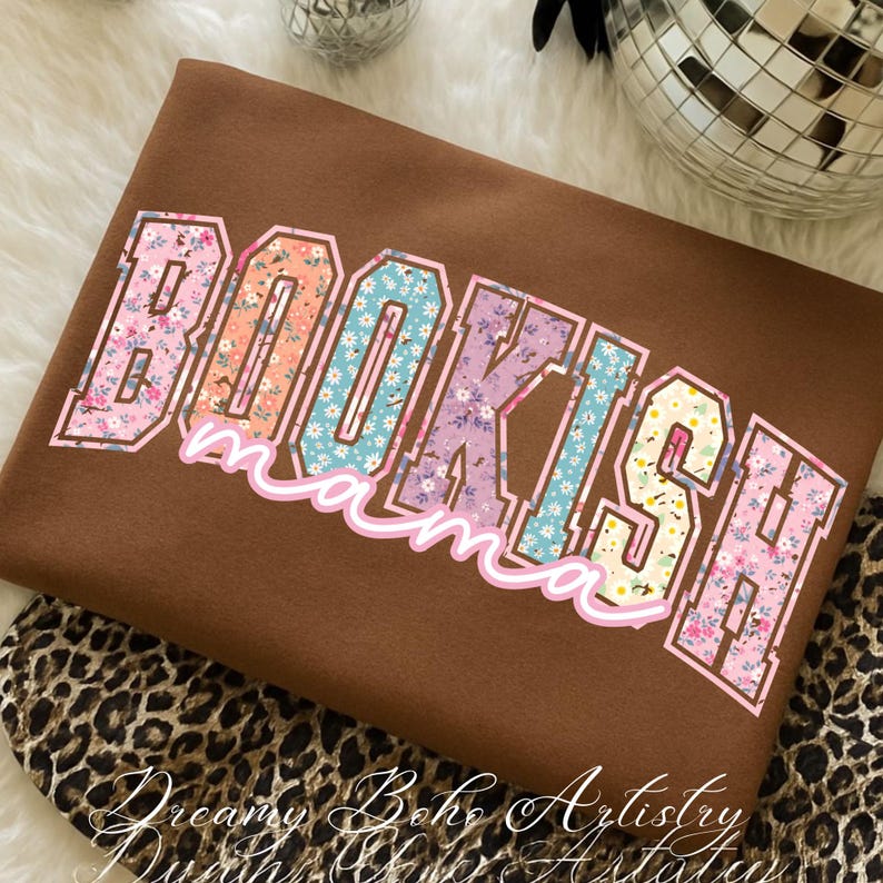 May include: A brown sweatshirt with the text "BOOKISH mama" in a floral design. The letters are outlined in white and filled with various floral patterns in pink, blue, orange, and purple. The sweatshirt is folded.