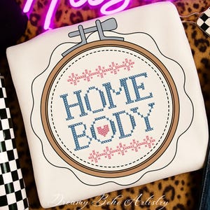 May include: A cream-coloured cushion with a cross-stitch design. The design features the words "HOME BODY" in blue, a pink heart, and decorative pink and blue cross-stitch elements within a brown embroidery hoop. The cushion has a scalloped edge.