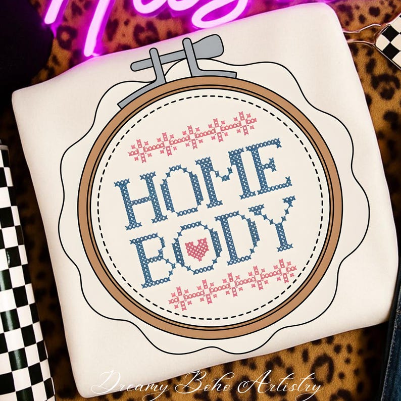 May include: A cream-coloured cushion with a cross-stitch design. The design features the words "HOME BODY" in blue, a pink heart, and decorative pink and blue cross-stitch elements within a brown embroidery hoop. The cushion has a scalloped edge.