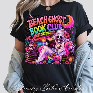 Puede incluir: Camiseta negra con un gr&aacute;fico colorido. Presenta el texto "BEACH GHOST BOOK CLUB" y una sirena leyendo un libro. El dise&ntilde;o incluye calaveras, un flamenco y una escena de puesta de sol. Tambi&eacute;n incluye el texto "reading from beyond the grave".