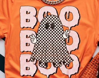 May include: Orange t-shirt with a graphic of a checkered ghost giving a peace sign, with the word "BOO" in pink bubble letters. The shirt has a crew neck and short sleeves. The text "Dreamy Boho Artistry" is at the bottom.
