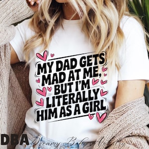 Może przedstawiać: Biała koszulka z czarnym napisem "MY DAD GETS MAD AT ME, BUT I'M LITERALLY HIM AS A GIRL" z różowymi serduszkami. Codzienna, casualowa odzież.
