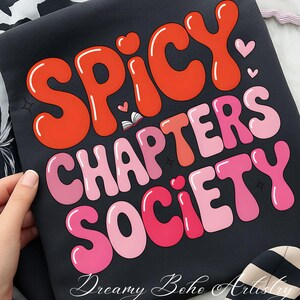 May include: Black fabric featuring the text "Spicy Chapters Society" in orange and pink bubble letters. Small hearts and a book graphic are included. The words "Dreamy Boho Artistry" are at the bottom of the fabric.