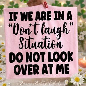 May include: A light pink t-shirt with the text "IF WE ARE IN A "Don't laugh" Situation DO NOT LOOK OVER AT ME" in black. The words "Dreamy Boho Artistry" are at the bottom. The shirt is held up by a person.