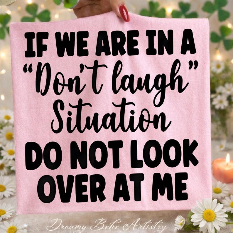 May include: A light pink t-shirt with the text "IF WE ARE IN A "Don't laugh" Situation DO NOT LOOK OVER AT ME" in black. The words "Dreamy Boho Artistry" are at the bottom. The shirt is held up by a person.