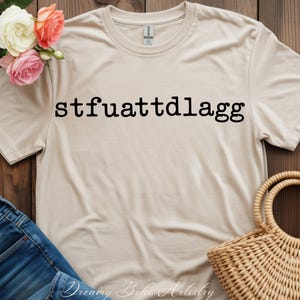 Puede incluir: Una camiseta beige con el texto "stfuattdlaggg" impreso en negro. La camiseta se muestra sobre una superficie de madera con unos vaqueros azules, una cesta de mimbre y un ramo de flores.