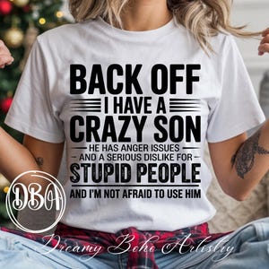 Könnte beinhalten: Weißes T-Shirt mit dem Text "BACK OFF I HAVE A CRAZY SON" in fetten schwarzen Buchstaben. Der Text geht weiter mit "HE HAS ANGER ISSUES AND A SERIOUS DISLIKE FOR STUPID PEOPLE AND I'M NOT AFRAID TO USE HIM". Das Shirt hat einen Rundhalsausschnitt.