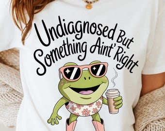May include: White t-shirt featuring a cartoon frog wearing pink sunglasses and cowboy boots, holding a coffee cup. The shirt has the text "Undiagnosed But Something Ain't Right" and "Dreamy Boho Artistry".