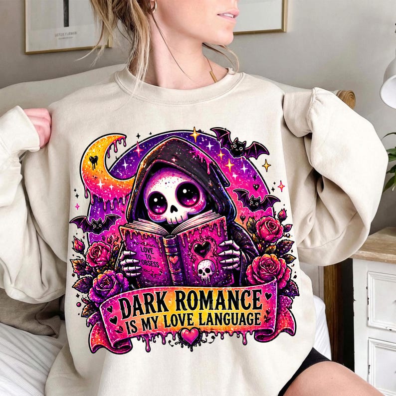 May include: Beige sweatshirt featuring a graphic of a hooded skeleton reading a book with the text "DARK ROMANCE IS MY LOVE LANGUAGE". The design includes bats, roses, a crescent moon, and stars in shades of purple, pink, and orange.