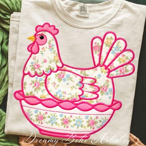 May include: A cream-colored t-shirt featuring a whimsical chicken design. The chicken is crafted from floral fabric with pink, blue, and yellow flowers, outlined in bright pink. The design includes a pink comb, beak, and wattle. The text "Dreamy Boho Artistry" is at the bottom.
