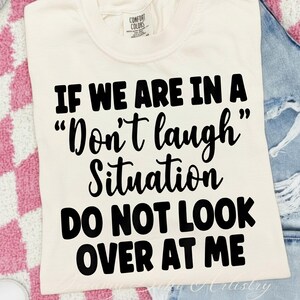 May include: Cream-colored t-shirt with the text "IF WE ARE IN A 'Don't laugh' Situation DO NOT LOOK OVER AT ME" in bold black letters. The shirt has a Comfort Colors tag in the upper left corner.