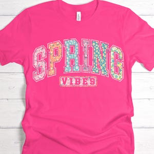 May include: A bright pink t-shirt with the words "SPRING VIBES" in a floral design. The word "SPRING" is arched across the top, and "VIBES" is in a rectangular box below.