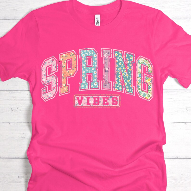 May include: A bright pink t-shirt with the words "SPRING VIBES" in a floral design. The word "SPRING" is arched across the top, and "VIBES" is in a rectangular box below.