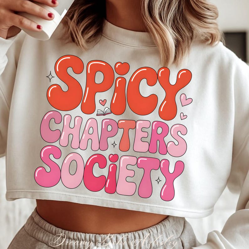 May include: Cream-coloured cropped sweatshirt featuring the text "SPICY CHAPTERS SOCIETY" in orange and pink bubble letters. The design includes small hearts, stars, and a book illustration. The sweatshirt has a crew neck.