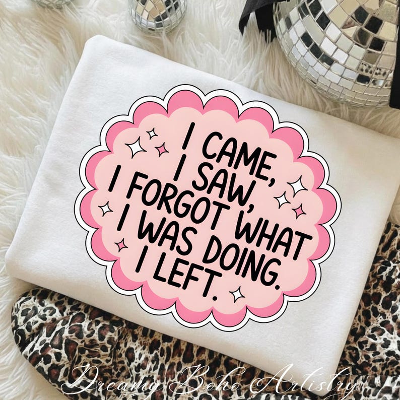 May include: White sweatshirt with a pink and white graphic that reads "I CAME, I SAW, I FORGOT WHAT I WAS DOING, I LEFT." The graphic is surrounded by a pink scalloped border and star accents. The sweatshirt is folded on top of a leopard print fabric.