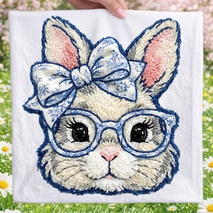 Könnte beinhalten: Weißes Stoffquadrat mit einem detaillierten, gestickten Hasengesicht. Der Hase trägt eine blau gerahmte Brille und eine passende Schleife mit floralem Muster. Das Fell ist eine Mischung aus Weiß, Grau und Beige, mit rosa Akzenten in Ohren und Nase.