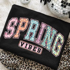 May include: Black sweatshirt with the word "SPRING" in a floral design, and the word "VIBES" in a rectangular box. The letters are outlined in white and the floral patterns include pink, orange, blue, and yellow.