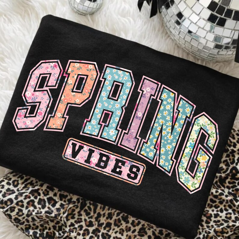 May include: Black sweatshirt with the word "SPRING" in a floral design, and the word "VIBES" in a rectangular box. The letters are outlined in white and the floral patterns include pink, orange, blue, and yellow.