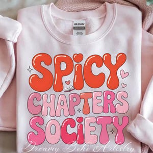May include: A pale pink Gildan sweatshirt featuring the text "SPICY CHAPTERS SOCIETY" in a retro bubble font. The words are in shades of orange and pink, with heart and star details. The sweatshirt is made from a heavy cotton blend.
