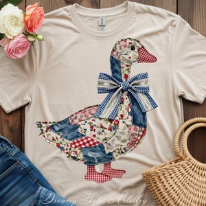 Peut inclure: T-shirt crème avec un motif d'oie en patchwork. L'oie est ornée de motifs floraux, denim et vichy, ainsi que d'un nœud bleu et blanc à carreaux. Le texte "Dreamy Boho Artistry" est en bas.