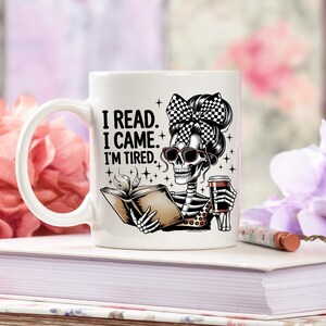 May include: White ceramic mug featuring a skeleton with sunglasses and a checkered bow, reading a book and holding a coffee cup. The mug has the text "I READ. I CAME. I'M TIRED." in black letters. The mug is on a stack of books.