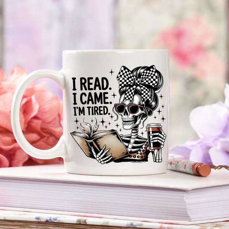 May include: White ceramic mug featuring a skeleton with sunglasses and a checkered bow, reading a book and holding a coffee cup. The mug has the text "I READ. I CAME. I'M TIRED." in black letters. The mug is on a stack of books.