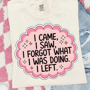 May include: Cream-colored t-shirt with a pink and white graphic that reads "I CAME, I SAW, I FORGOT WHAT I WAS DOING. I LEFT." The graphic has a scalloped edge and star accents. The shirt has a Comfort Colors tag.