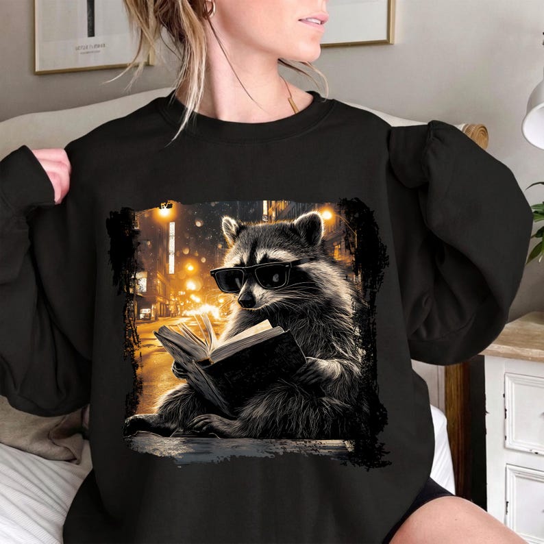 May include: Black sweatshirt featuring a raccoon wearing sunglasses and reading a book. The raccoon is set against a backdrop of a city street at night, with warm yellow lights. The graphic is framed by a black border.