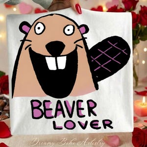 Puede incluir: Camiseta blanca con un diseño de castor de dibujos animados. El castor tiene una cara marrón, boca negra y cola negra. Las palabras "BEAVER LOVER" están impresas en rosa y negro. De Dreamy Boho Artistry.