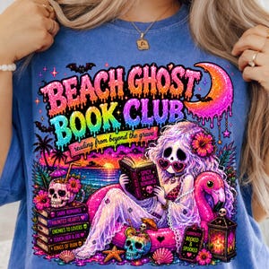Puede incluir: Camiseta azul con un gr&aacute;fico colorido que presenta el texto "BEACH GHOST BOOK CLUB" y una sirena leyendo un libro. El dise&ntilde;o incluye calaveras, un flamenco y las palabras "reading from beyond the grave".