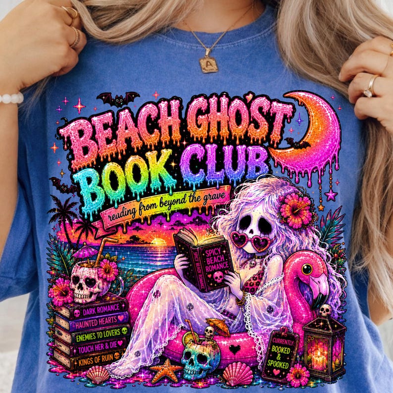 Puede incluir: Camiseta azul con un gr&aacute;fico colorido que presenta el texto "BEACH GHOST BOOK CLUB" y una sirena leyendo un libro. El dise&ntilde;o incluye calaveras, un flamenco y las palabras "reading from beyond the grave".