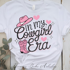 Może przedstawiać: Biała koszulka z napisem "In my Cowgirl Era" w czarnym skrypcie, ozdobiona różowymi sercami, różowym kapeluszem kowbojskim i różowymi kowbojkami. Koszulka ma okrągły dekolt i krótkie rękawy.