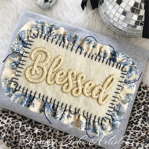 May include: A gray textile with a cream-colored rectangular patch embroidered with the word "Blessed" in gold thread. The patch is bordered with blue floral fabric and dark blue stitching. The words "Dreamy Boho Artistry" are at the bottom.