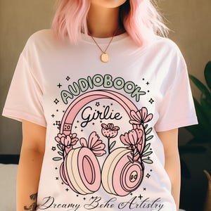 May include: White t-shirt with a pink and green graphic design of headphones with flowers and the text "Audiobook Girlie" and "Dreamy Boho Artistry".