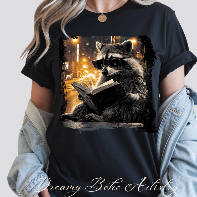 May include: Black t-shirt featuring a raccoon wearing sunglasses and reading a book. The raccoon is set against a blurred cityscape with warm, golden streetlights. The text "Dreamy Boho Artistry" is at the bottom.