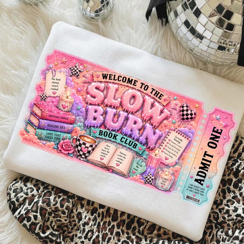 May include: White sweatshirt with a colorful graphic that reads "Welcome to the Slow Burn Book Club." The design includes books, a coffee cup, candles, and the words "Admit One." The sweatshirt is decorated with pink, purple, and blue hues.