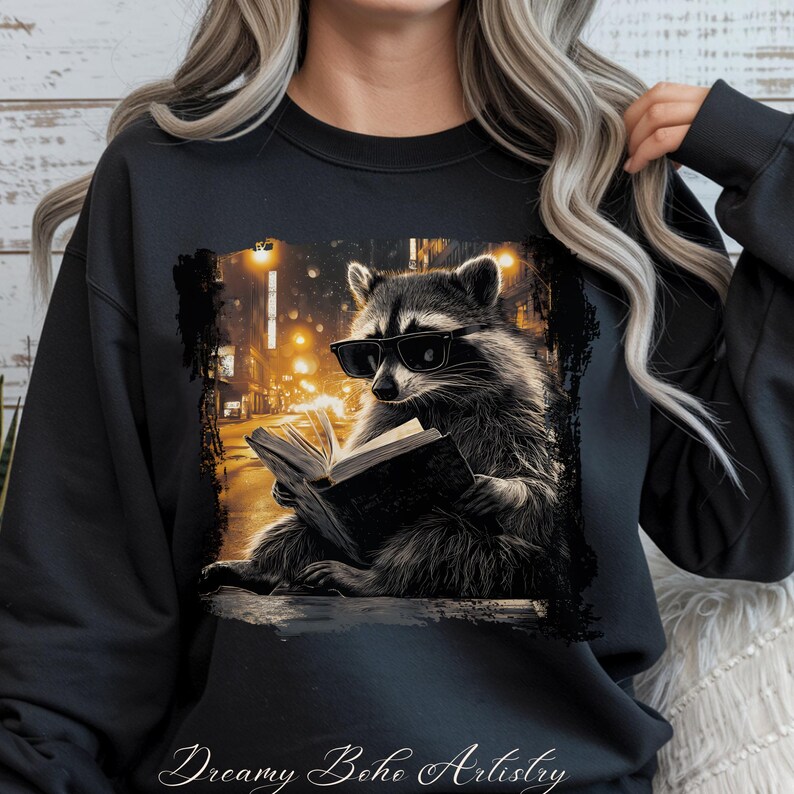 May include: Black sweatshirt featuring a raccoon wearing sunglasses and reading a book. The raccoon is set against a blurred city street scene with warm, yellow streetlights. The words "Dreamy Boho Artistry" are at the bottom.