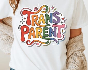 May include: White t-shirt with a colorful graphic that reads "TRANS PARENT" in rainbow colors. The design has a retro, sticker-like aesthetic with a white outline and star accents. The shirt is part of a clothing collection.