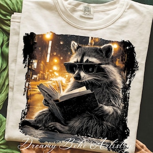 May include: Cream-colored t-shirt featuring a raccoon wearing sunglasses and reading a book. The raccoon is set against a blurred cityscape with warm, golden streetlights. The graphic is framed by a black border.