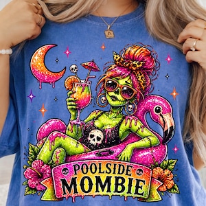 May include: Blue t-shirt featuring a green zombie woman with sunglasses, holding a cocktail while floating on a pink flamingo pool float. The shirt has the text "Poolside Mombie" on a banner with floral accents.