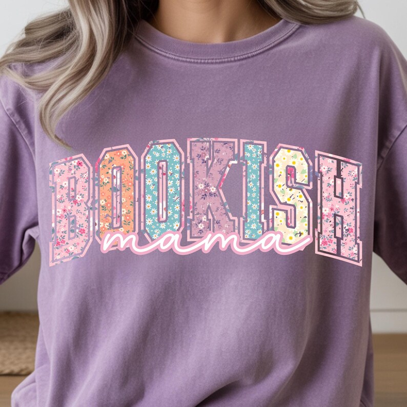 May include: A purple sweatshirt with the text "BOOKISH mama" in a floral and colorful design. The word "BOOKISH" is in a block letter style with a floral pattern, and "mama" is in a cursive font.