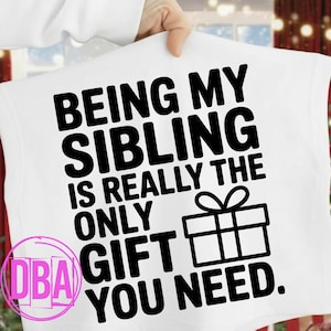 May include: White t-shirt with the text "BEING MY SIBLING IS REALLY THE ONLY GIFT YOU NEED" in bold black letters. A black and white gift box graphic is also present. The shirt is held up by a person.
