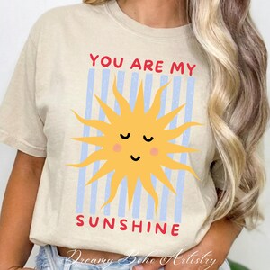 May include: Beige t-shirt with a yellow sun graphic and the text "YOU ARE MY SUNSHINE" in red. The sun has a smiling face and is set against blue vertical stripes. The shirt is a casual, everyday wear item.