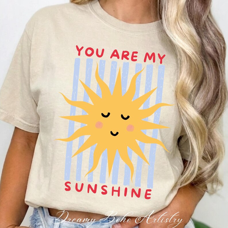 May include: Beige t-shirt with a yellow sun graphic and the text "YOU ARE MY SUNSHINE" in red. The sun has a smiling face and is set against blue vertical stripes. The shirt is a casual, everyday wear item.